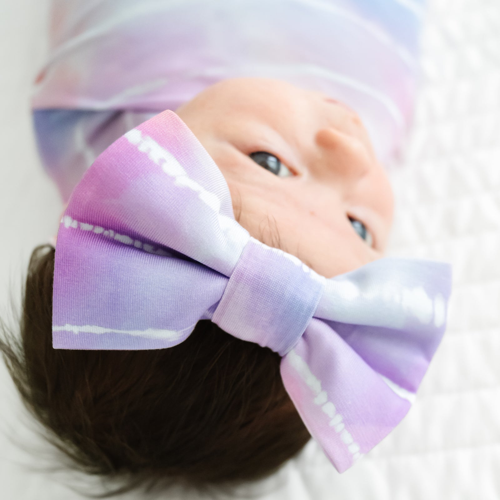 Close up image of an infant swaddled in a Pastel Tie Dye Dreams swaddle & luxe bow headband set, detailing the luxe bow headband