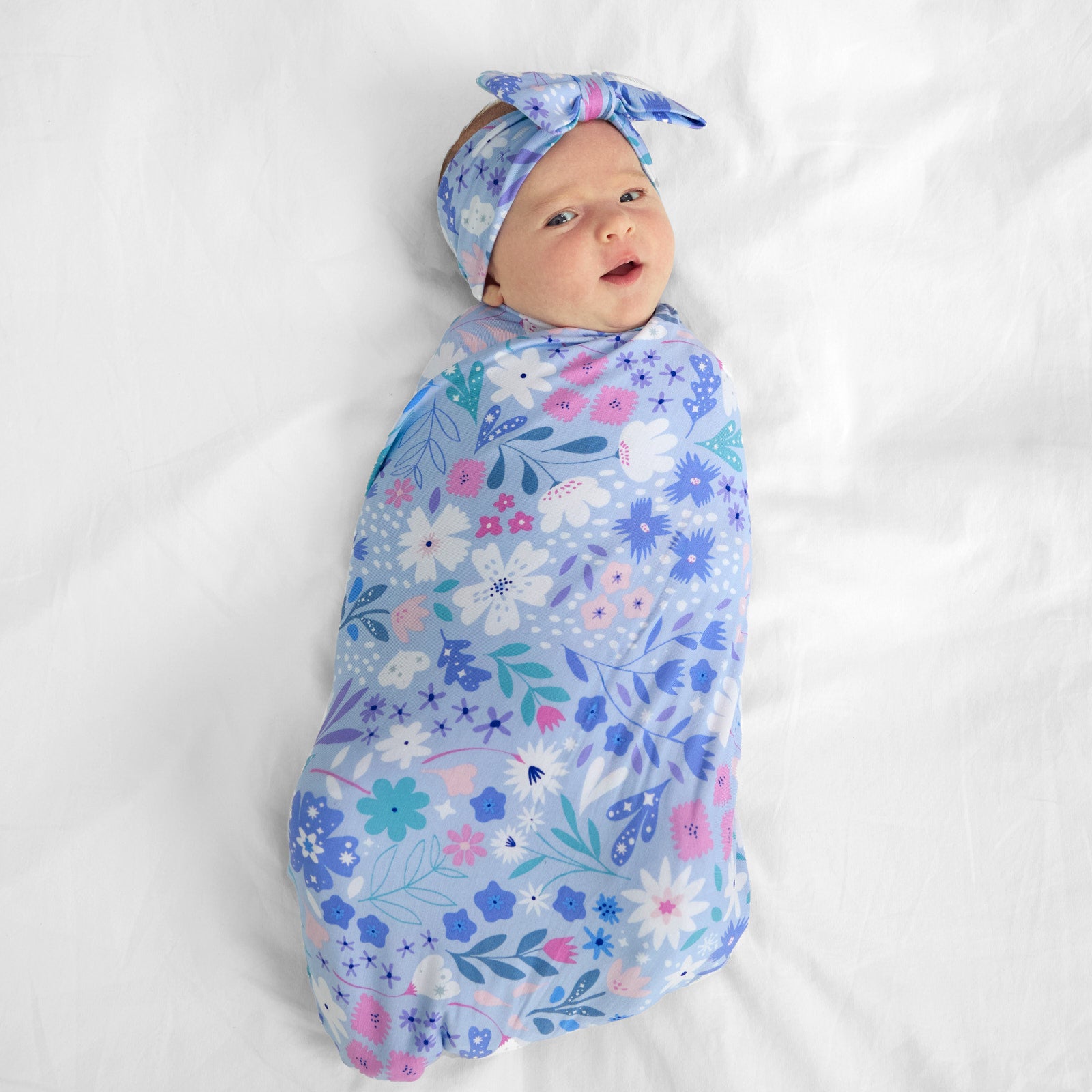 Image of a child swaddled on a bed swaddled in a Magical Meadow swaddle and luxe bow headband set