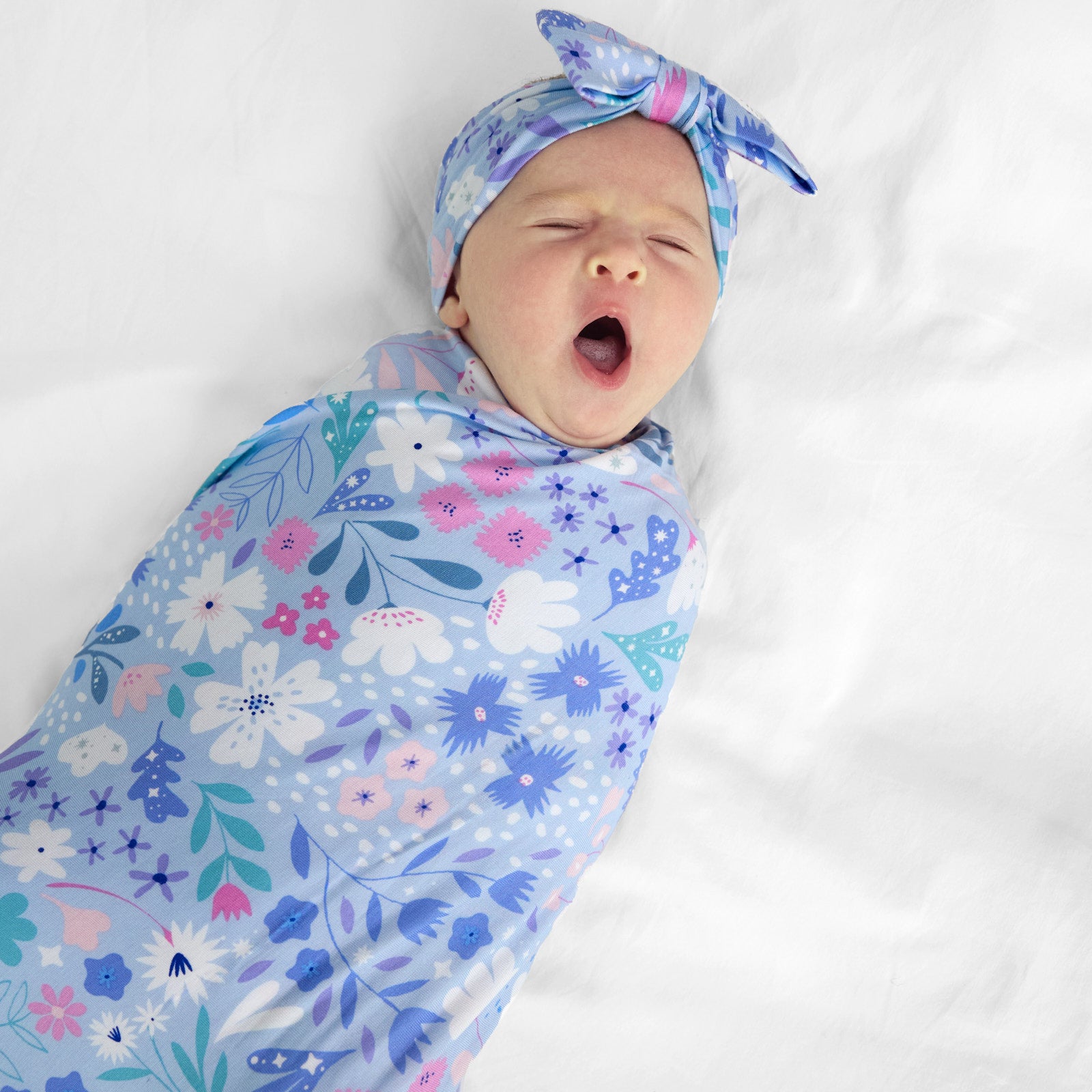 Close up image of a child swaddled on a bed swaddled in a Magical Meadow swaddle and luxe bow headband set