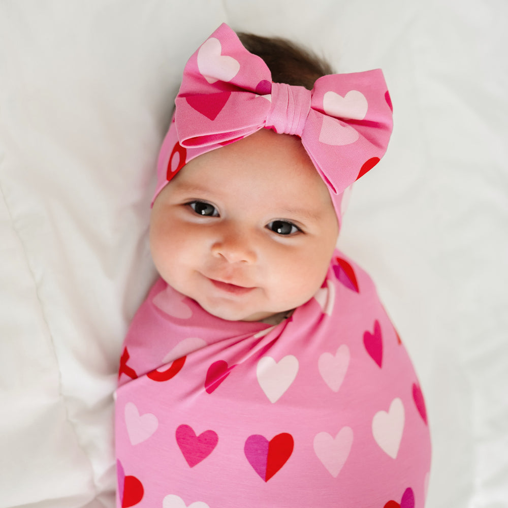Click to see full screen - Close up image of a child laying on a bed swaddled in a Pink XOXO swaddle and luxe bow set