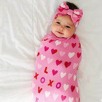 Child laying on a bed swaddled in a Pink XOXO swaddle and luxe bow set