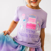 Close up image of a child wearing a Magical Birthday Light Lavender graphic tee paired with a rainbow tutu skirt