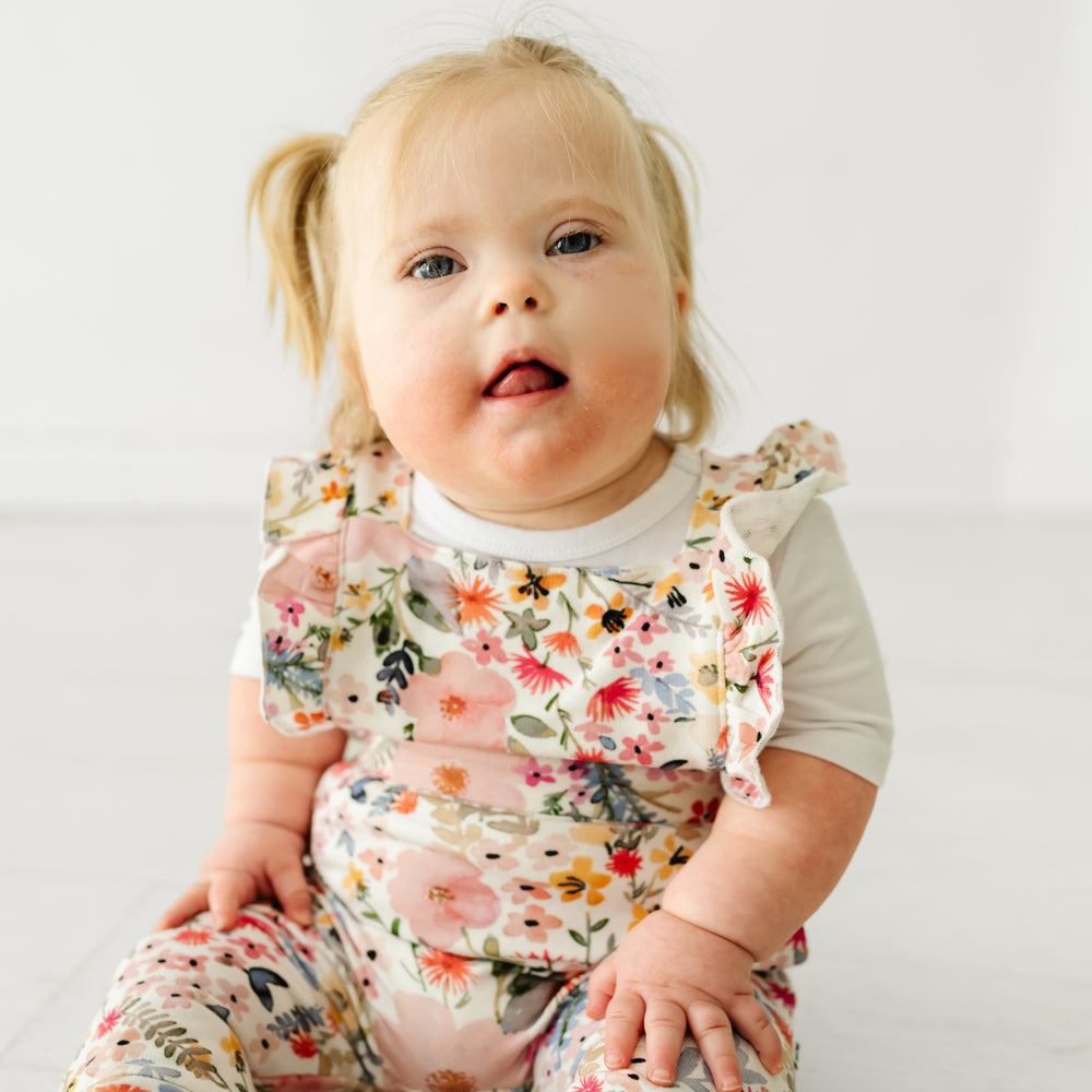 Child sitting on the ground wearing a Mauve Meadow ruffle overall and coordinating Play top