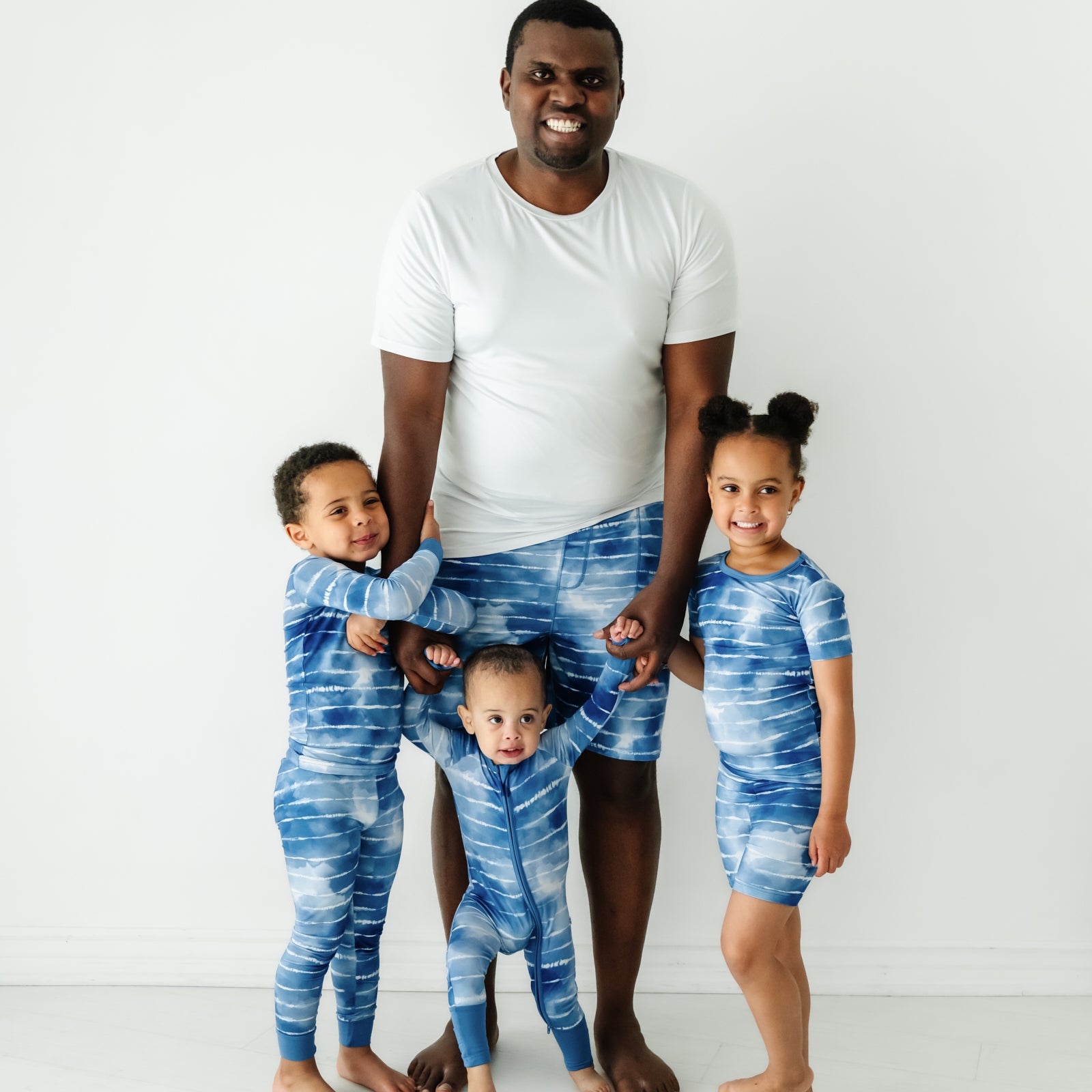 Dad and his three children wearing matching pjs. Dad is wearing a men's bright white pajama top paired with men's Blue Tie Dye Dreams pj shorts. Children are wearing Blue Tie Dye Dreams pajamas in two piece and zippy styles