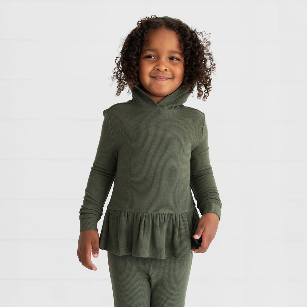 Close up image of a child wearing an Olive Waffle Peplum Hooded Tee with the hood up and matching Flare Pants