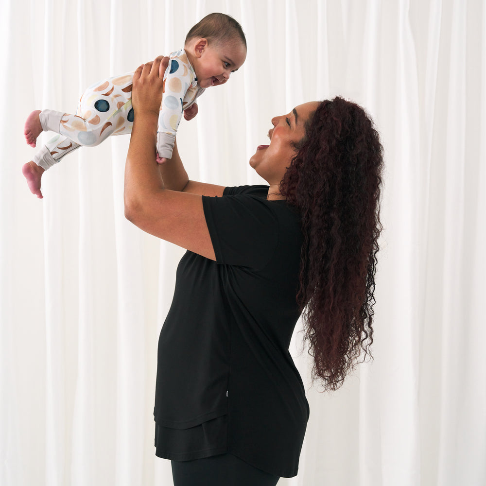 profile view of a woman wearing a Black women's nursing top holding up her child wearing a Luna Neutral zippy