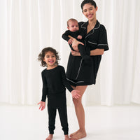 Image of a woman with her two children. Mom is wearing a solid Black women's short sleeve and shorts pajama set. Her kids are wearing Solid Black pjs in two piece and zippy styles