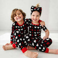Two children sitting together wearing matching Black XOXO two piece pajama sets in long and short sleeve styles paired with a matching luxe bow headband