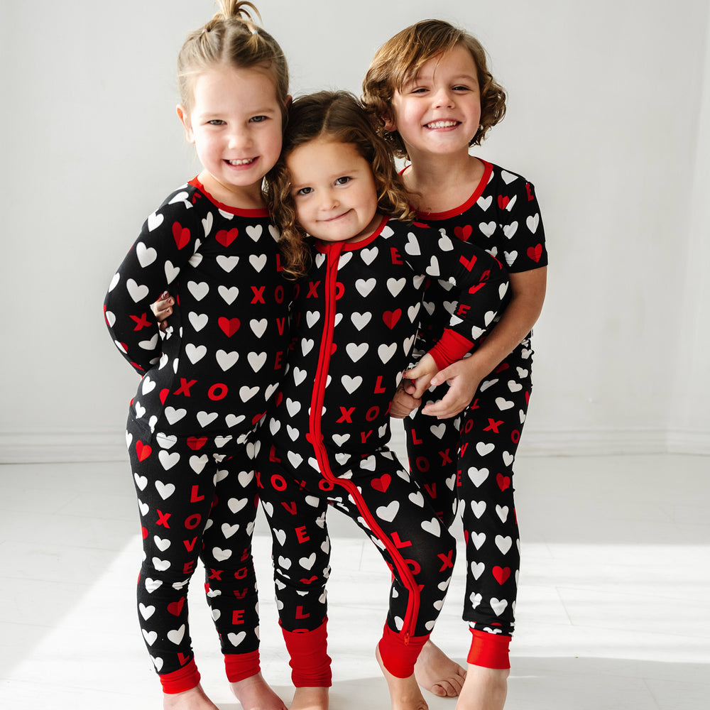 Click to see full screen - Three children holding hands wearing matching Black XOXO pajama sets. One is wearing a Black XOXO zippy and the other two are wearing Black XOXO pjs in long sleeve and short sleeve two piece styles
