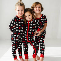 Three children holding hands wearing matching Black XOXO pajama sets. One is wearing a Black XOXO zippy and the other two are wearing Black XOXO pjs in long sleeve and short sleeve two piece styles