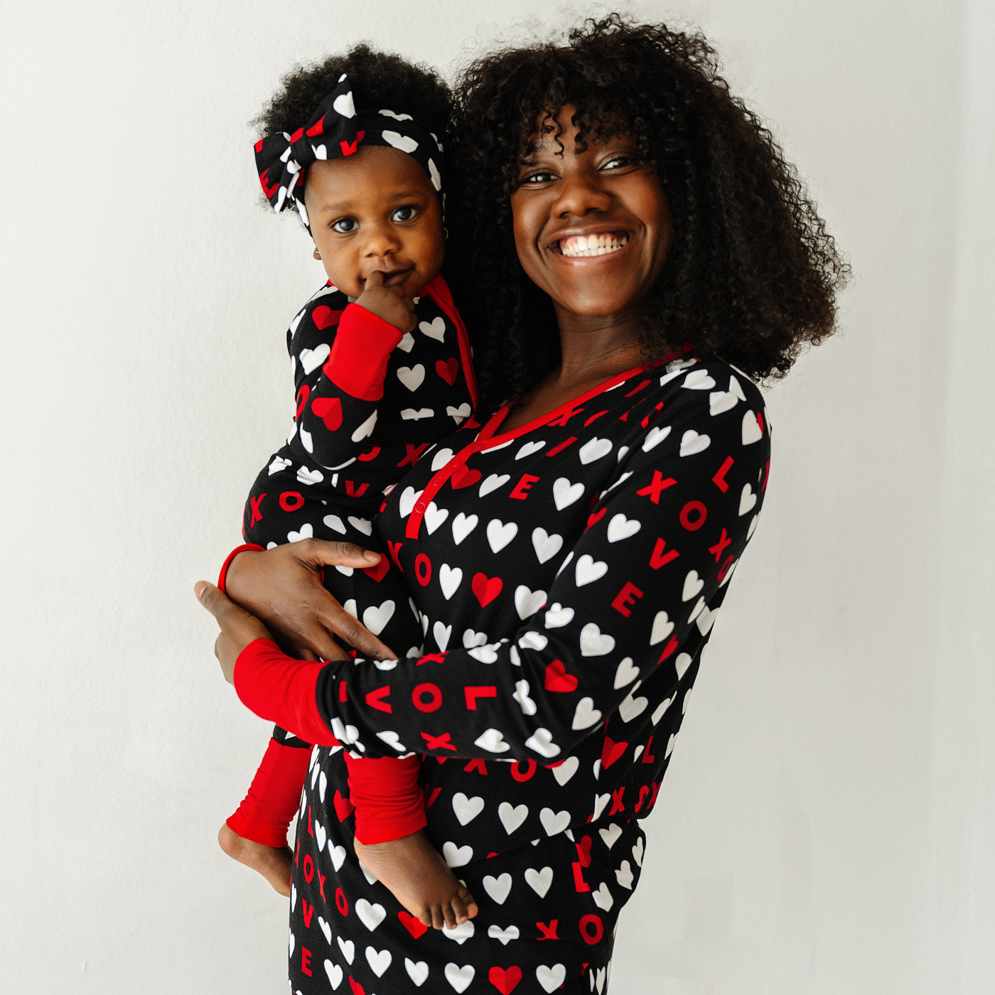 Mom holding her daughter wearing matching Black XOXO pajama sets. Mom is wearing a Black XOXO women's pajama top and child is wearing a Black XOXO zippy paired with a matching luxe bow headband.