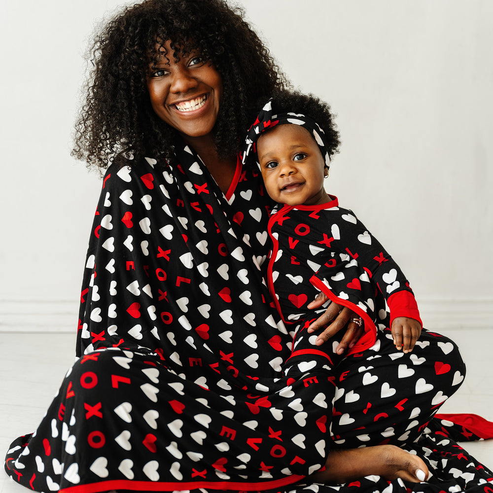 Click to see full screen - Mother and daughter wrapped up in a Black XOXO Oversized Cloud Blanket and wearing matching Black XOXO pajamas. Mom is wearing women's Black XOXO pajama top and matching pajama bottoms. Her daughter is wearing a Black XOXO zippy paired with a matching luxe bow headband