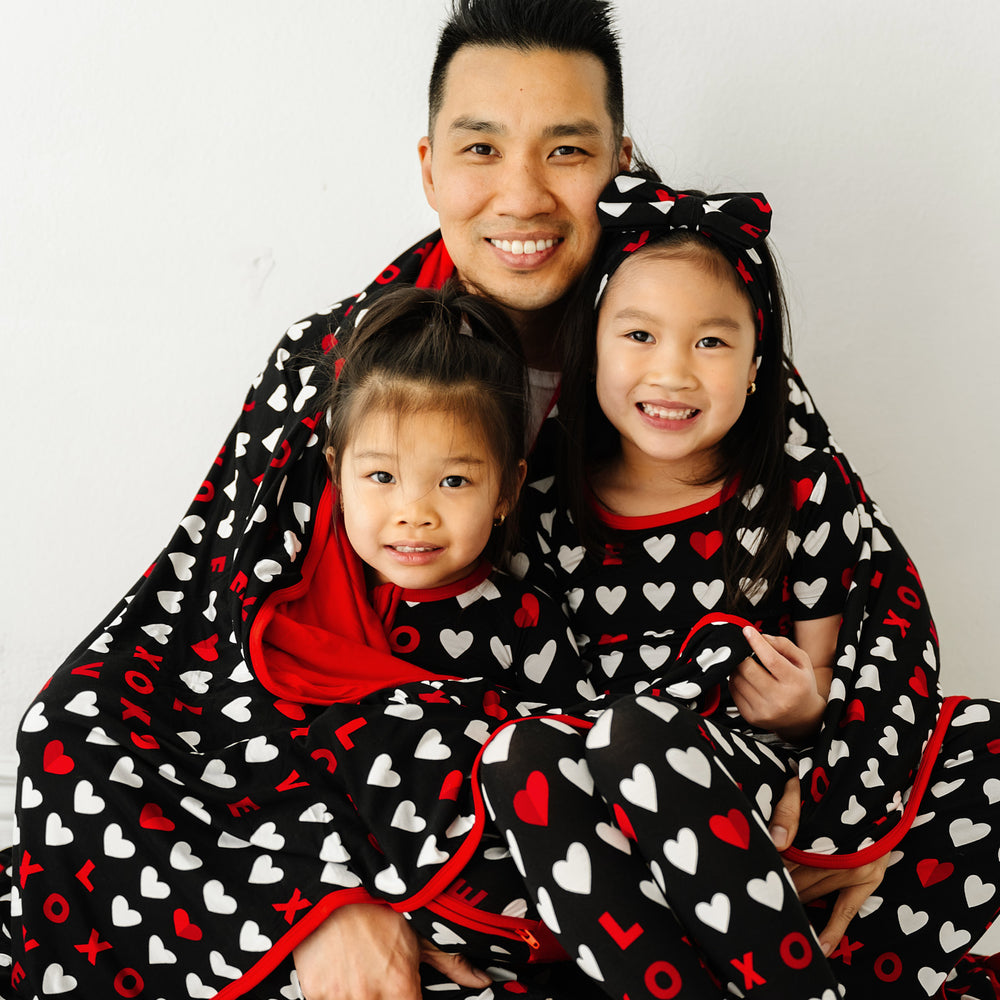 Click to see full screen - Family of three wrapped in a Black XOXO Oversized Cloud Blanket. Children are wearing matching Black XOXO pajama sets paired with a matching luxe bow headband