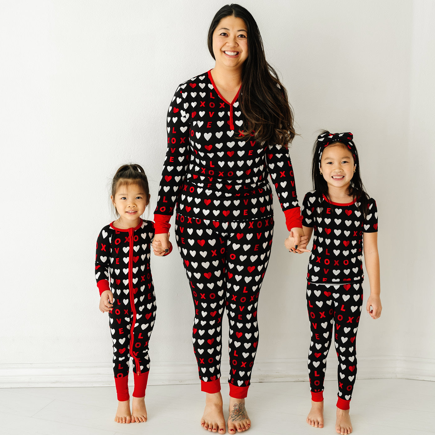 Mother and her two daughters holding hands wearing matching Black XOXO pajama sets. Mom is wearing a women's Black XOXO pajama top and matching women's pajama pants. Children are wearing Black XOXO pajamas in zippy and short sleeve two piece styles paired with a  matching luxe bow headband