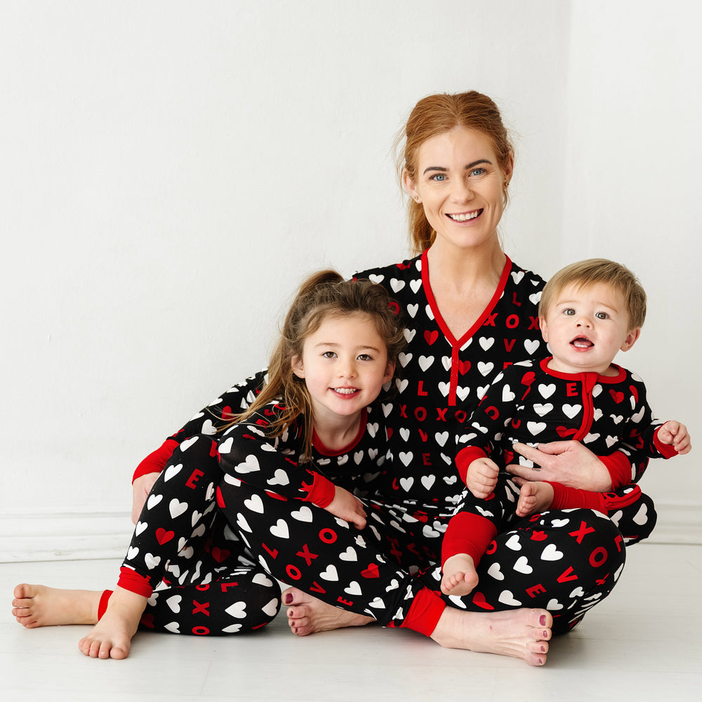 Click to see full screen - Mother and her two children sitting together wearing matching Black XOXO pajama sets. Mom is wearing a women's Black XOXO pajama top and matching women's pajama pants. Children are wearing Black XOXO pajamas in zippy and two piece styles