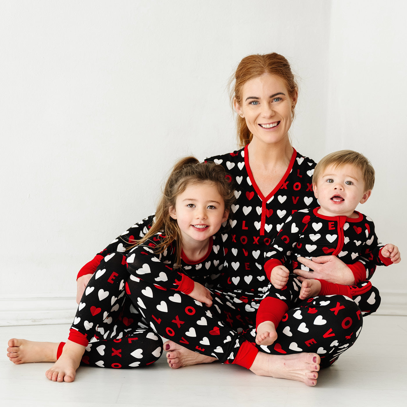 Mother and her two children sitting together wearing matching Black XOXO pajama sets. Mom is wearing a women's Black XOXO pajama top and matching women's pajama pants. Children are wearing Black XOXO pajamas in zippy and two piece styles