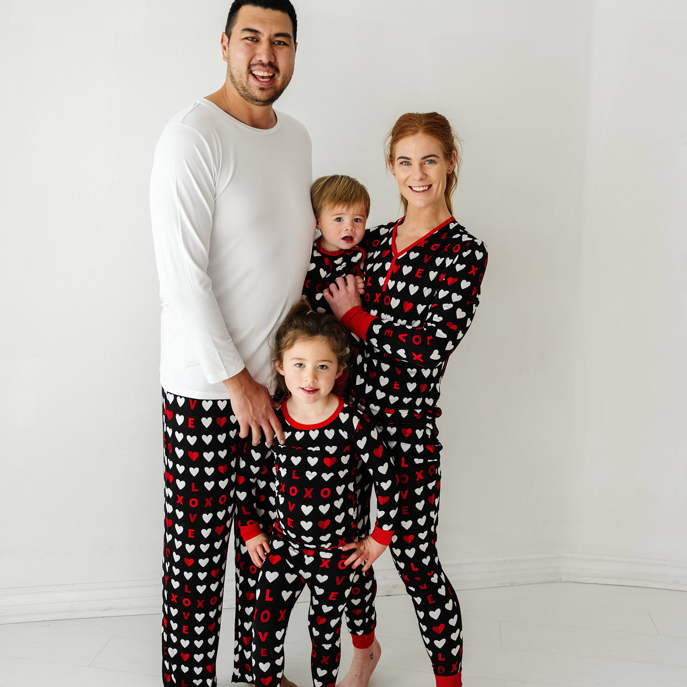 Family of four wearing matching Black XOXO pajama sets. Dad is wearing men's Black XOXO pajama pants paired with a men's solid white pajama top. Mom is wearing Black XOXO women's pajama pants and matching women's pajama top. Kids are wearing Black XOXO pajamas in two piece and zippy styles.