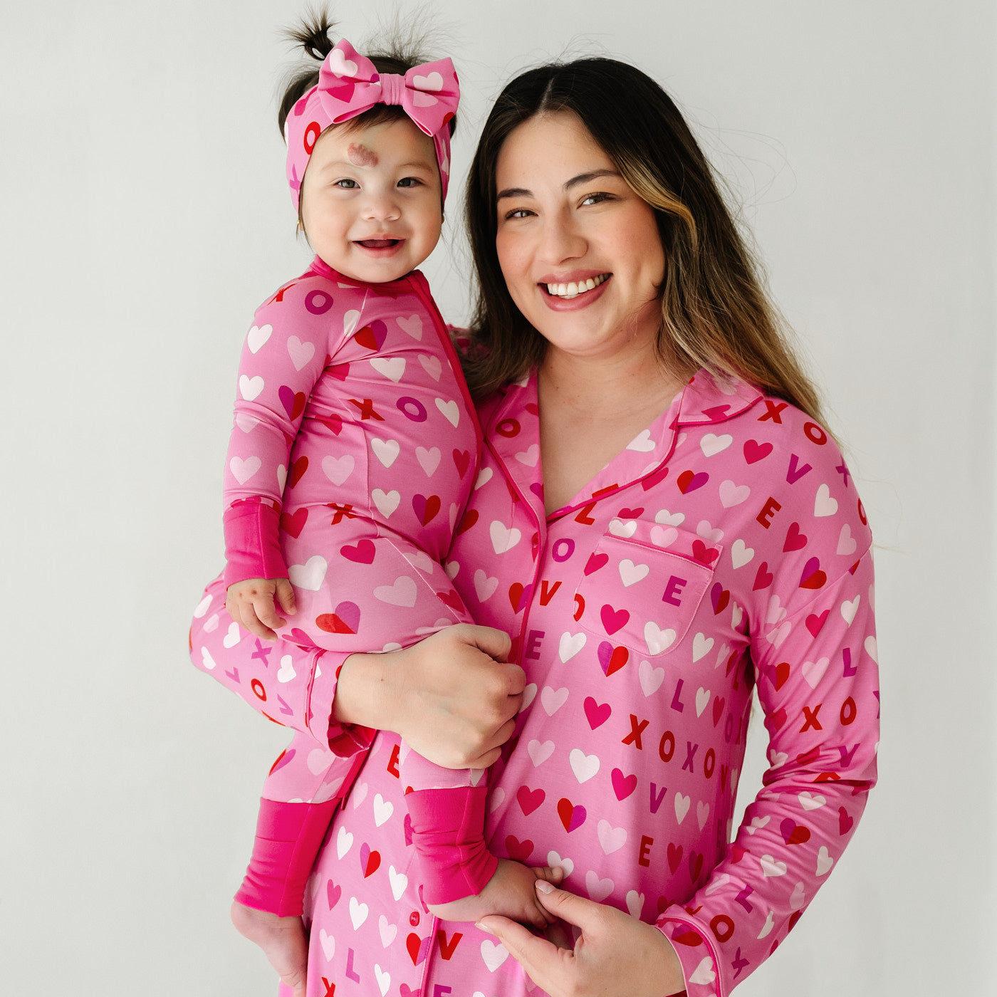 Mom holding her child wearing matching Pink XOXO pajamas. Child is wearing a Pink XOXO zippy paired with a matching luxe bow headband. Mom is wearing a women's Pink XOXO long sleeve sleep shirt