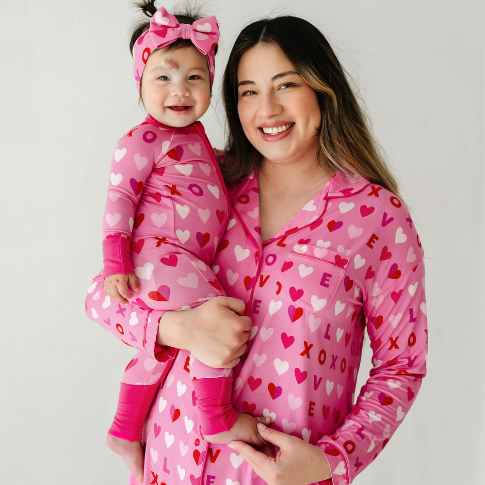 Click to see full screen - Mom holding her child wearing matching Pink XOXO pajamas. Child is wearing a Pink XOXO zippy paired with a matching luxe bow headband. Mom is wearing a women's Pink XOXO long sleeve sleep shirt