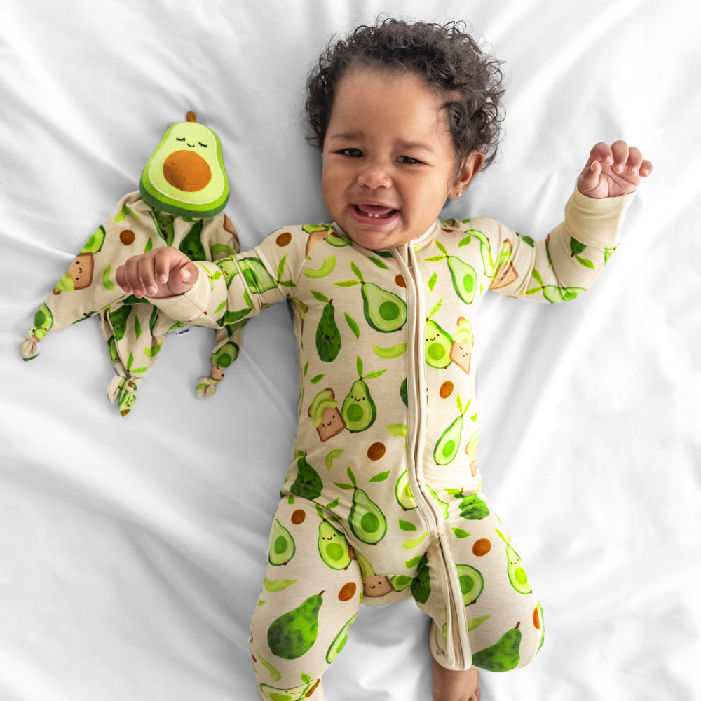 Image of a child wearing an Avocado Toast zippy on a bed with a matching Guac the Avocado Sleepyhead Lovey