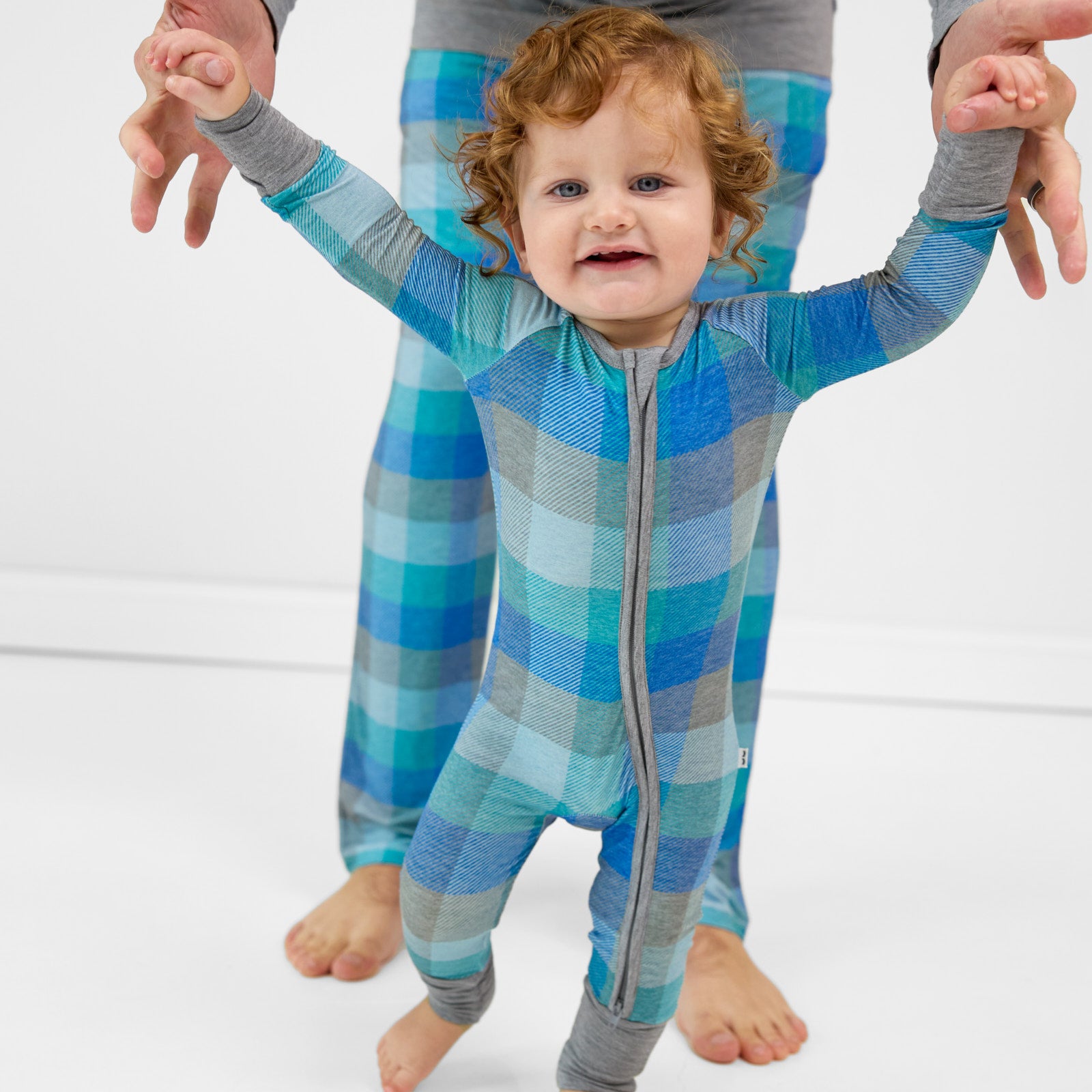 Child walking with help from their father. Child is wearing an Arctic Plaid zippy and their father is wearing matching Arctic Plaid men's pj pants