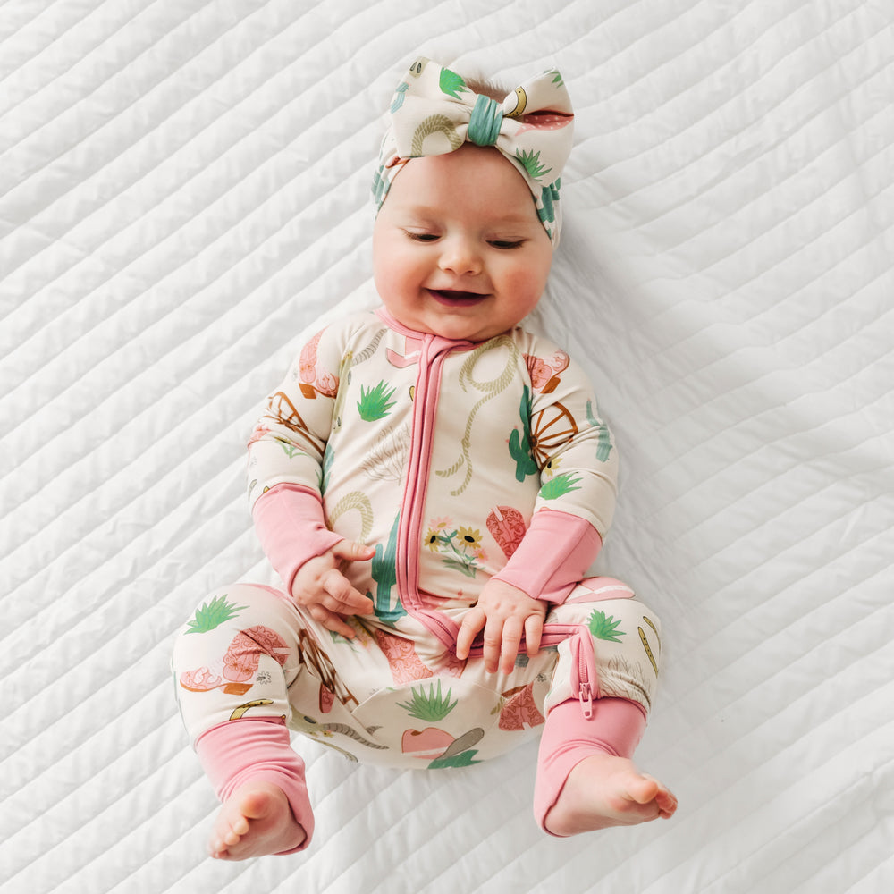 Child laying on a bed wearing a pink Ready to Rodeo zippy paired with a matching luxe bow headband