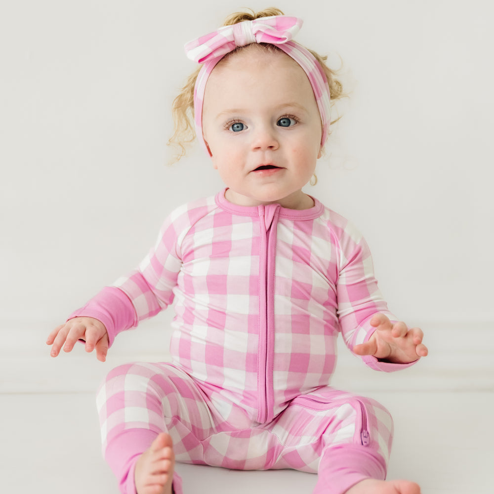 Click to see full screen - Child sitting wearing a Pink Gingham zippy paired with a matching luxe bow headband