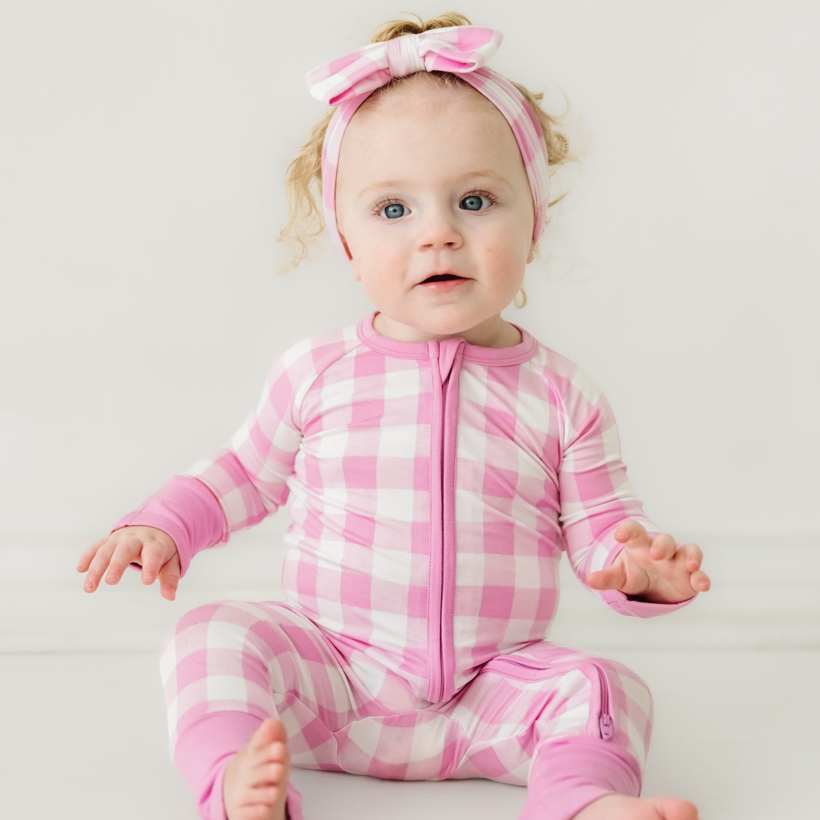 Child sitting wearing a Pink Gingham zippy paired with a matching luxe bow headband