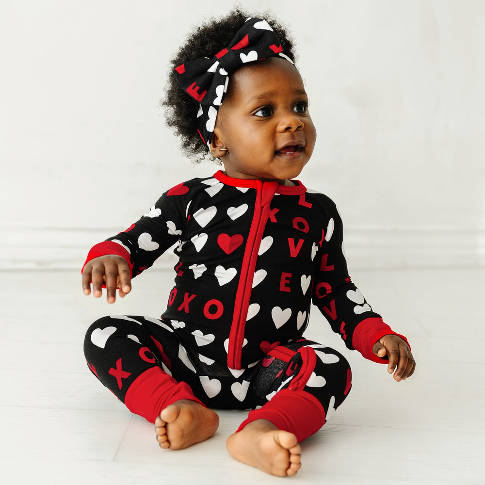 Click to see full screen - Image of a child sitting wearing a Black XOXO zippy paired with a matching luxe bow headband