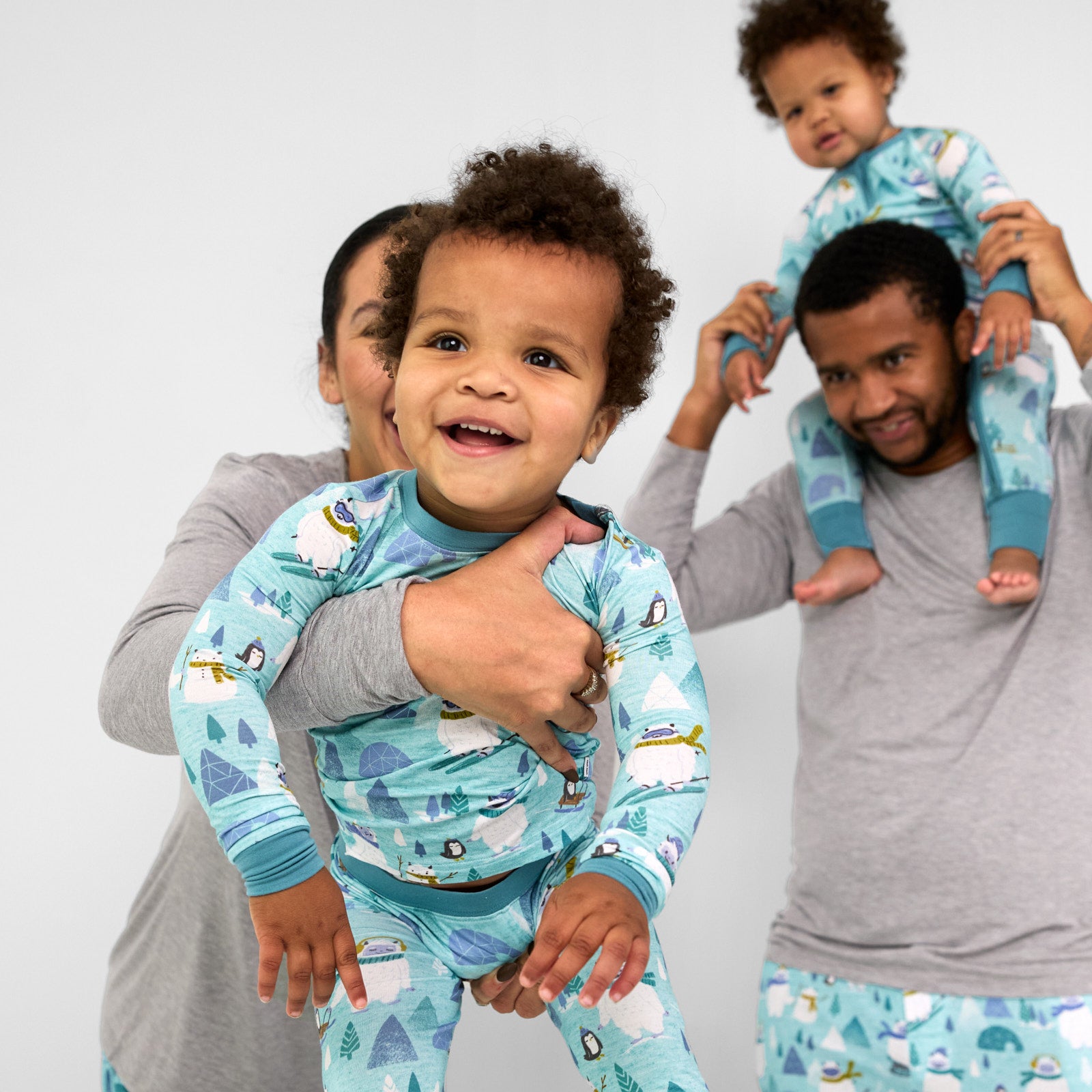 Alternate family of four wearing matching Yeti Snow Day pjs. Mom is wearing women's Heather Gray women's pj top and Yeti Snow Day women's pj pants. Dad is wearing a men's Heather Gray men's pj top and men's Yeti Snow Day pj pants. Their children are wearing a Yeti Snow Day zippy and two piece pj set. Mom is holding her child wearing a two piece set out in front of the camera