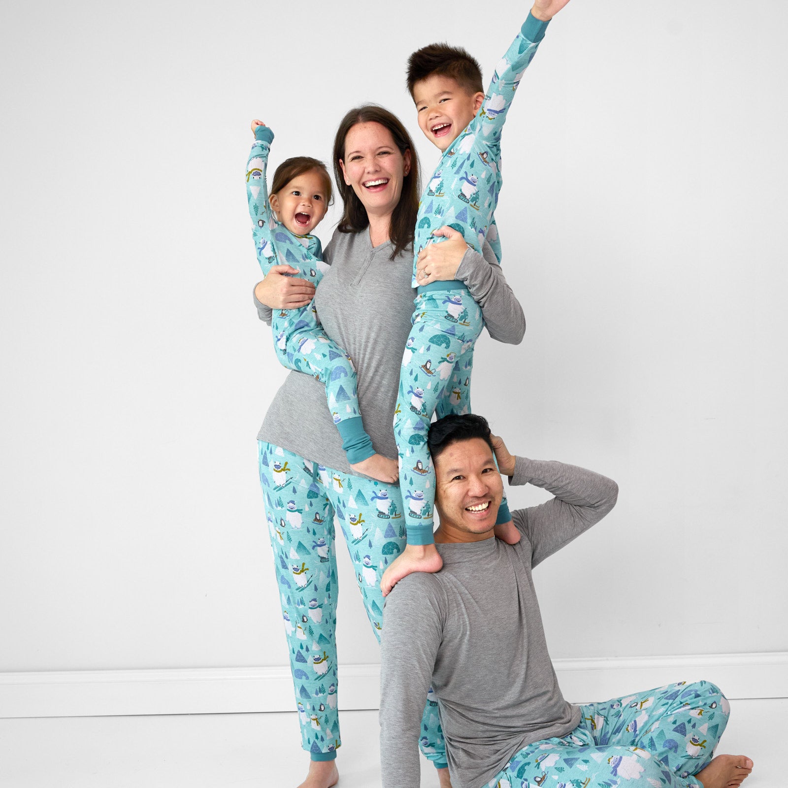 Family of four wearing matching Yeti Snow Day pjs. Mom is wearing women's Heather Gray women's pj top and Yeti Snow Day women's pj pants. Dad is wearing a men's Heather Gray men's pj top and men's Yeti Snow Day pj pants. Their children are wearing a Yeti Snow Day zippy and two piece pj set