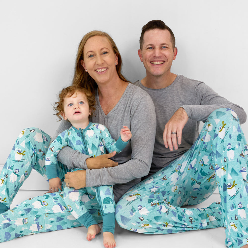 Family of three wearing matching Yeti Snow Day pjs. Mom is wearing women's Heather Gray women's pj top and Yeti Snow Day women's pj pants. Dad is wearing a men's Heather Gray men's pj top and men's Yeti Snow Day pj pants. Their child is wearing a Yeti Snow Day zippy