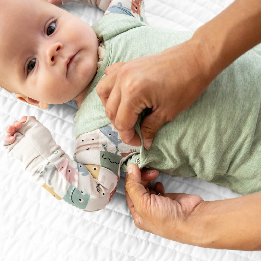 Child laying on a bed wearing a Heather Sage Sleepy bag over a Check Mates zippy. Mom is demonstrating how the underarm snaps work.