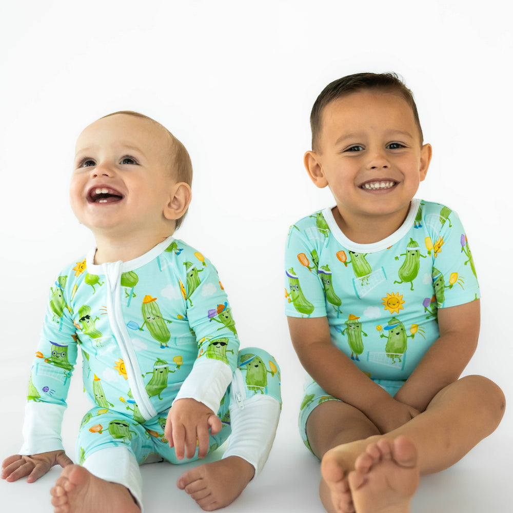 Two children sitting while wearing the Pickle Power print. Baby on the left is wearing the Pickle Power Zippy and boy on the right is wearing Pickle Power Two-Piece Short Sleeve & Shorts Pajama Set