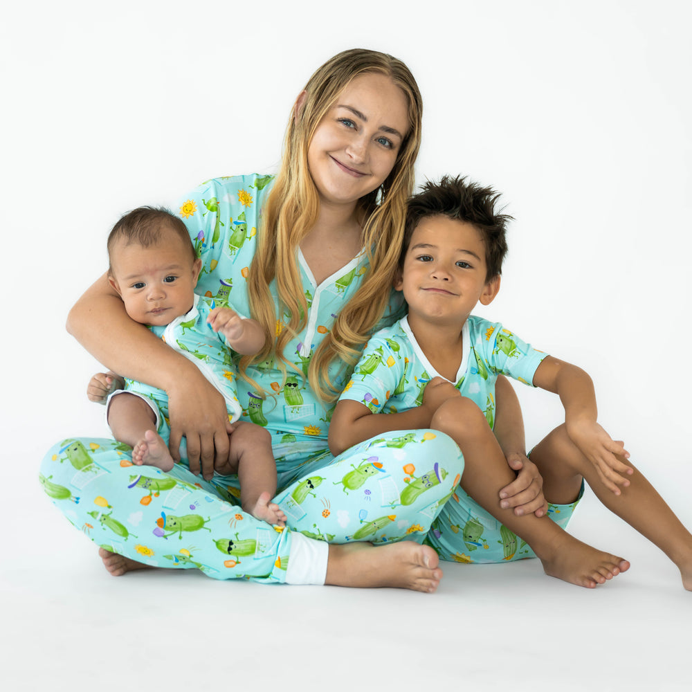 Family sitting while wearing the Pickle Power print. Baby on the left is wearing the Pickle Power Shorty Zippy and mother in the middle is wearing the Pickle Power Women's Pajama Pants and Short-sleeve Pajama Top. Boy on the left is wearing the Pickle Power Short-sleeve Shorts Pajama Set