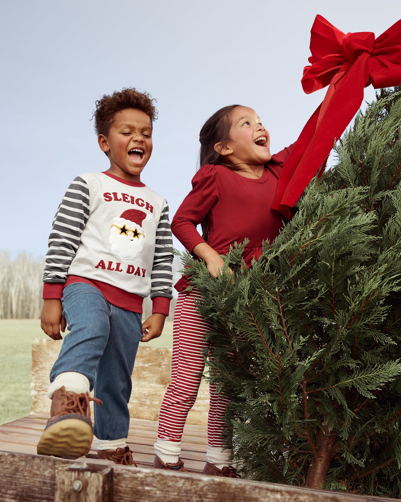 Two kids at a Christmas tree farm wearing styles from the Holiday Play Collection