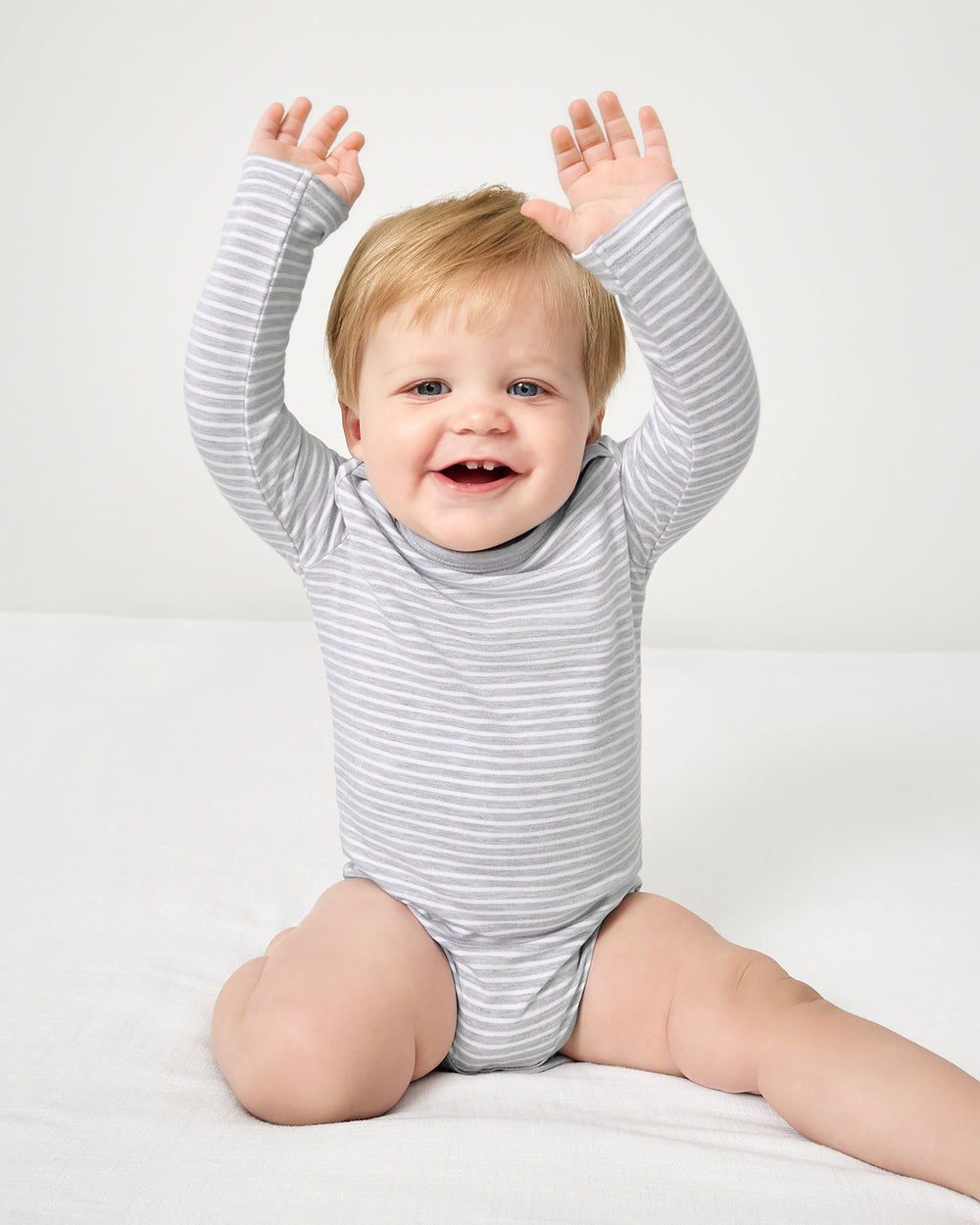 Baby sitting on a white bed wearing the Light Heather Gray Stripe Bodysuit