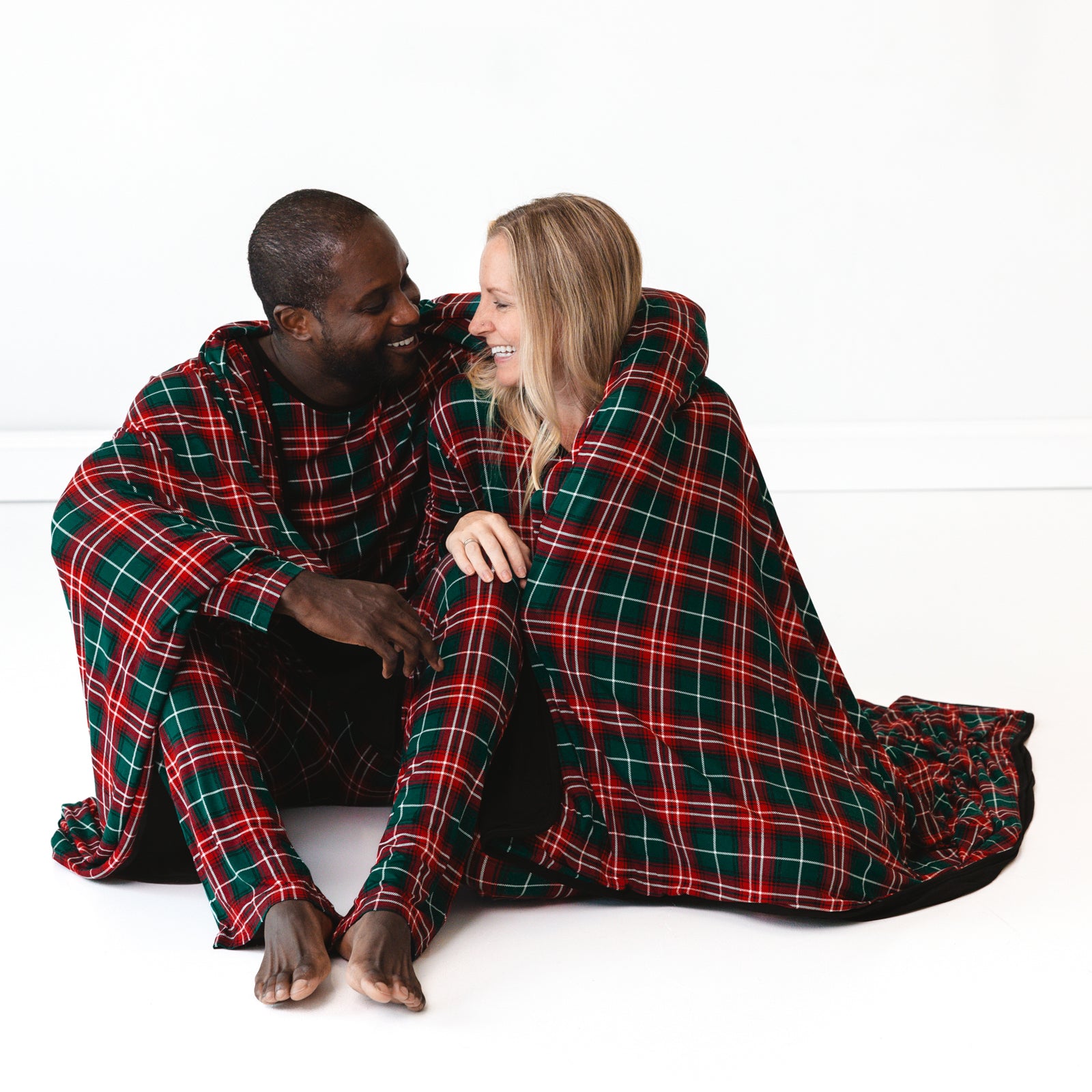 Two parents sitting on the ground with a Fireside Plaid Oversized Cloud Blanket® wrapped around them. They are wearing matching Fireside Plaid pajamas