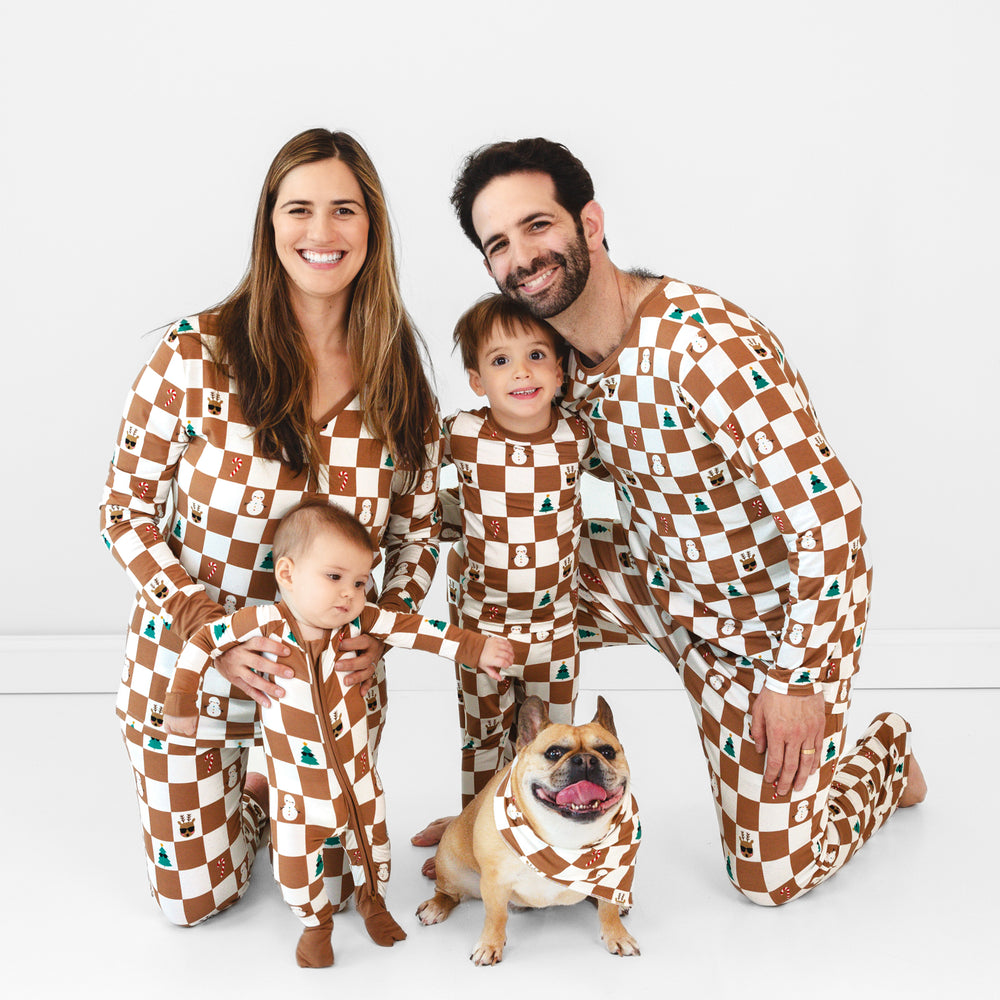 Family of four plus dog wearing the Holiday Checks print. Female model on the left is wearing the Holiday Checks Women's Pajama Top and Pants while holding standing baby in the Holiday Checks Zippy. Child in the middle is wearing the Holiday Checks Two-piece Pajama Set while hugging ale model who is wearing the Holiday Checks Men's Pajama Pants and Top. Dog in the middle in the Holiday Checks Pet Bandana