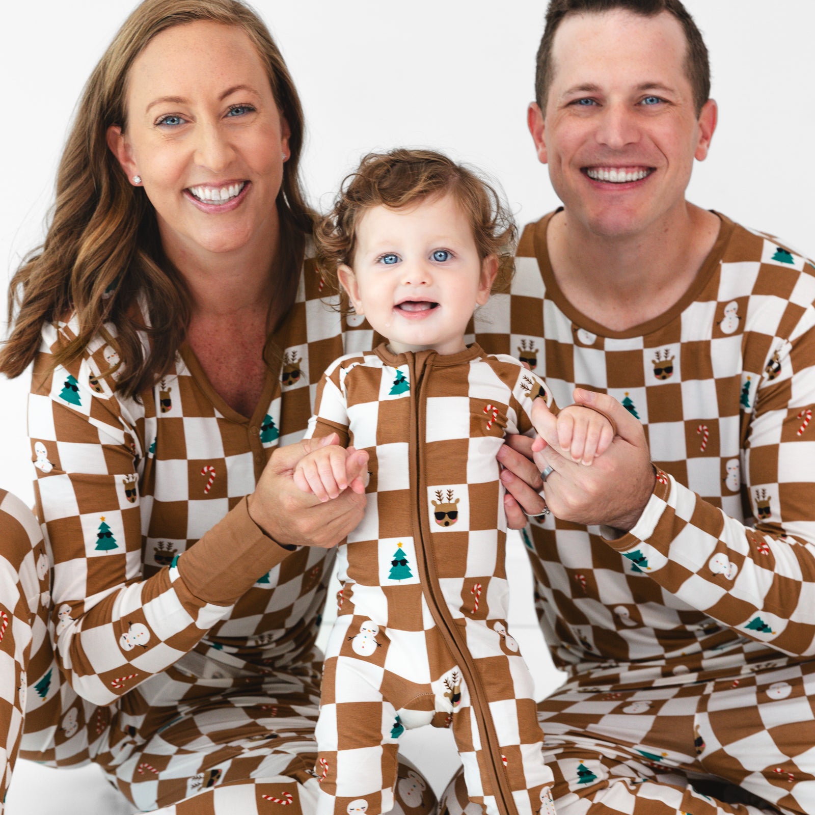 Smiling family wearing the Holiday Checks print. Female model is wearing the Holiday Checks Women's Pajama Top and Pants. Baby in the middle is wearing the. Holiday Checks Zippy and male model is wearing the Holiday Checks Men's Pajama Top and Pants