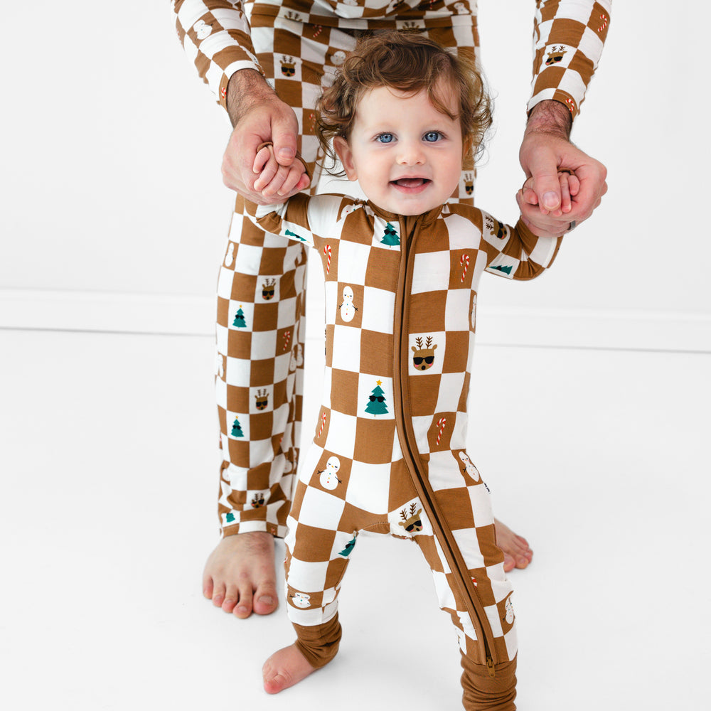 Baby standing while wearing the Holiday Checks Zippy, with help from male model who in the Holiday Checks Men's Pajama Pants