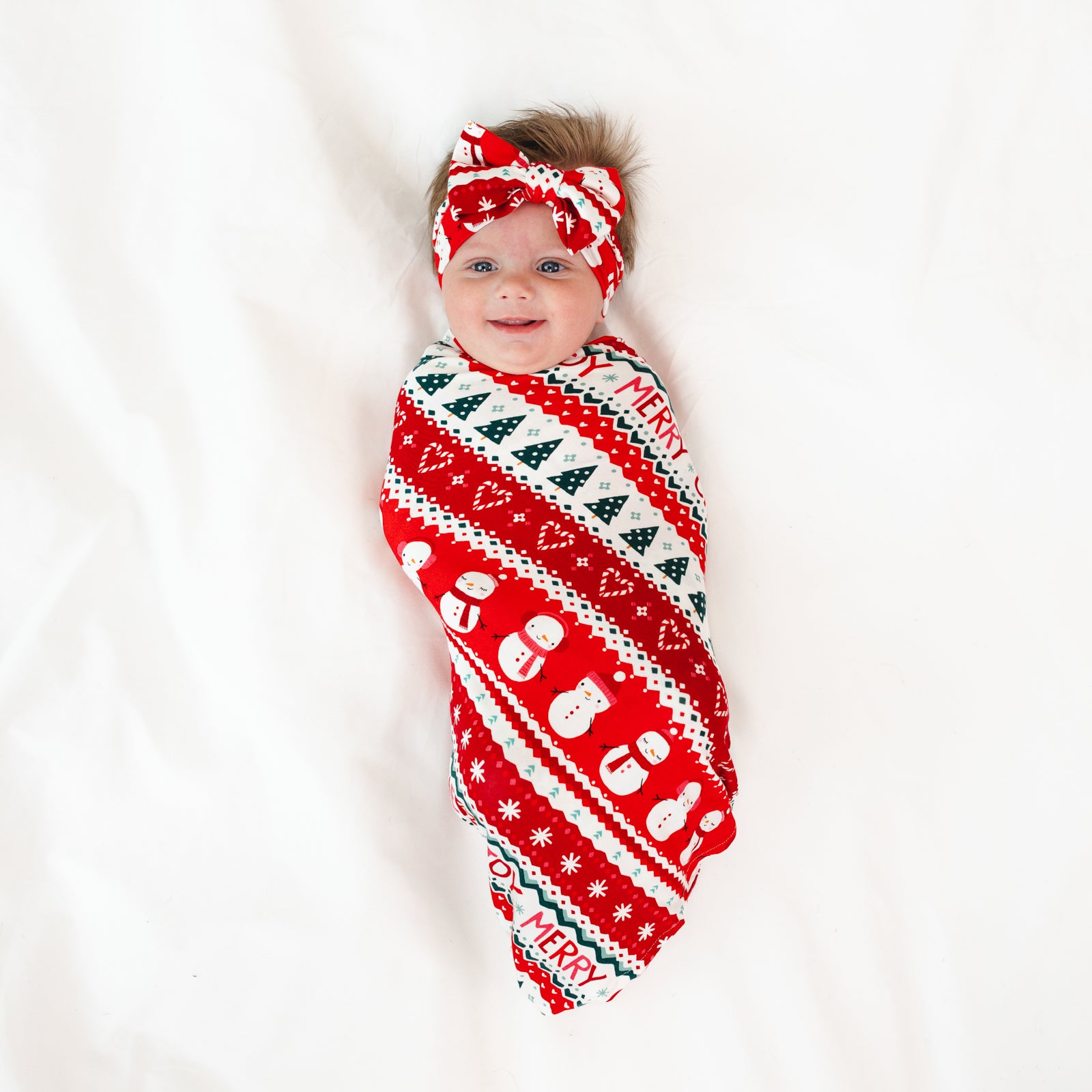 Image of a child laying on a blanket swaddled in a Festive Fair Isle Swaddle & Luxe Bow Headband Set