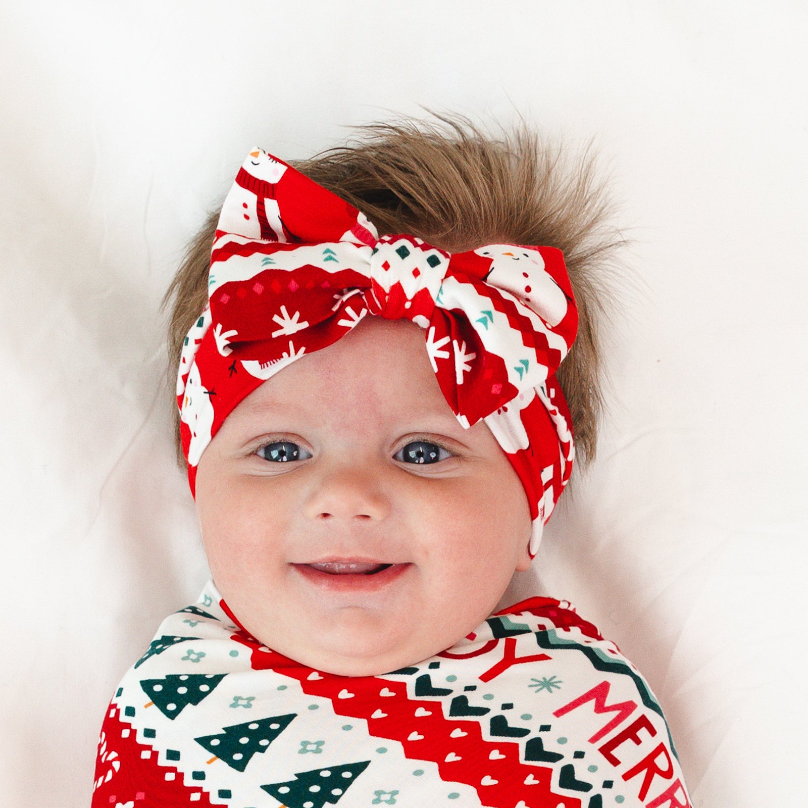 Close up image of a child laying on a blanket swaddled in a Festive Fair Isle Swaddle & Luxe Bow Headband Set