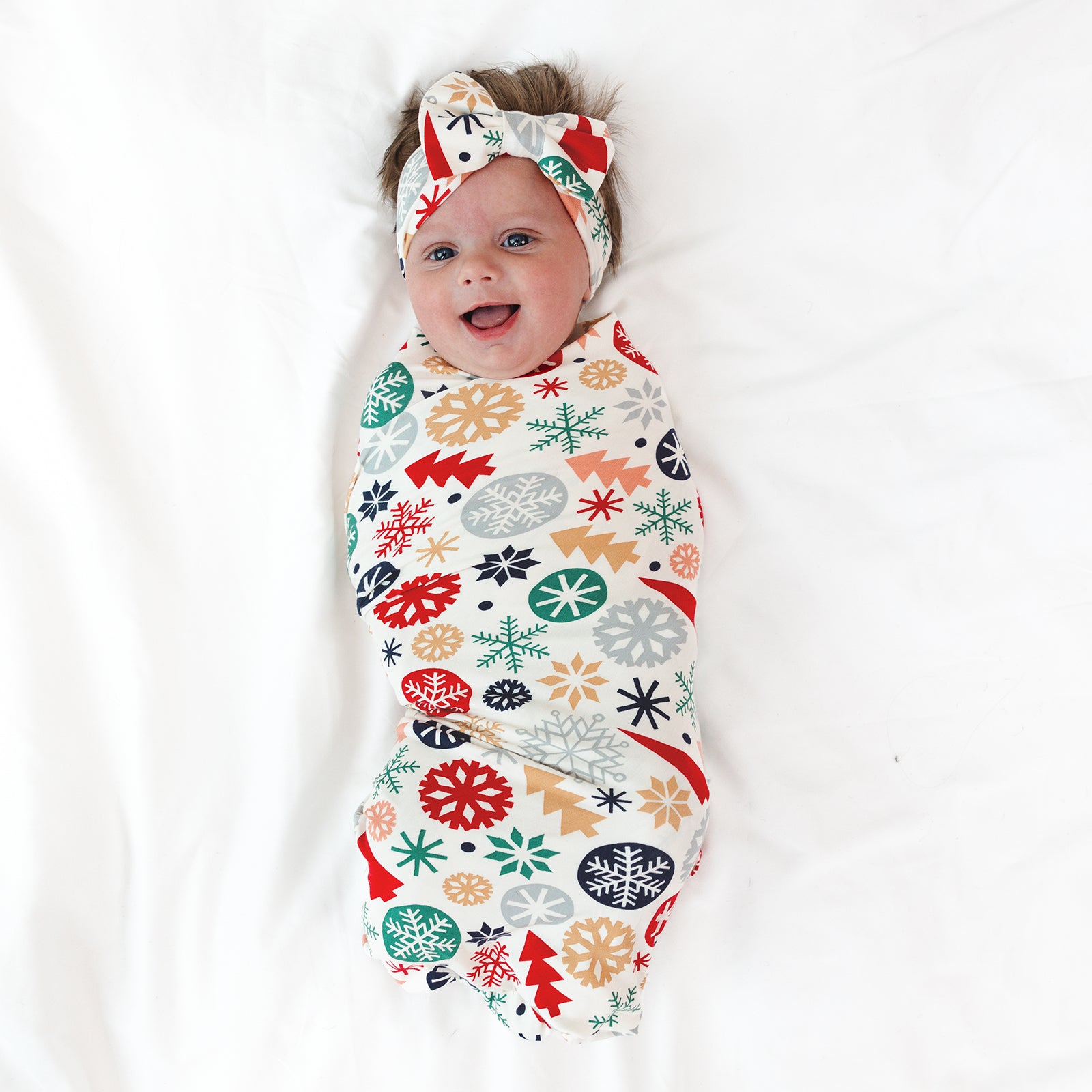 Top view image of smiling baby laying down wearing the Modern & Merry Swaddle & Luxe Bow Headband Set