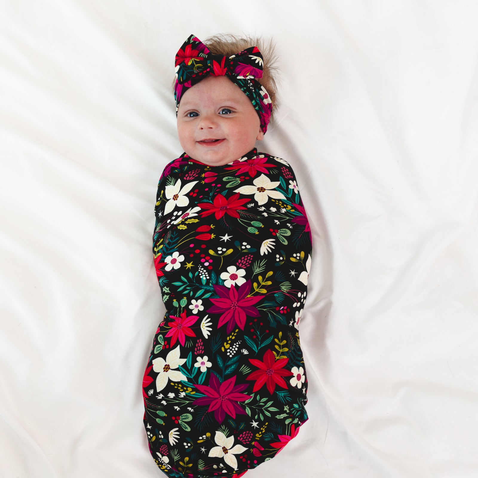 Top view image of baby laying down and smiling while in the Berry Merry Swaddle & Luxe Bow Headband Set