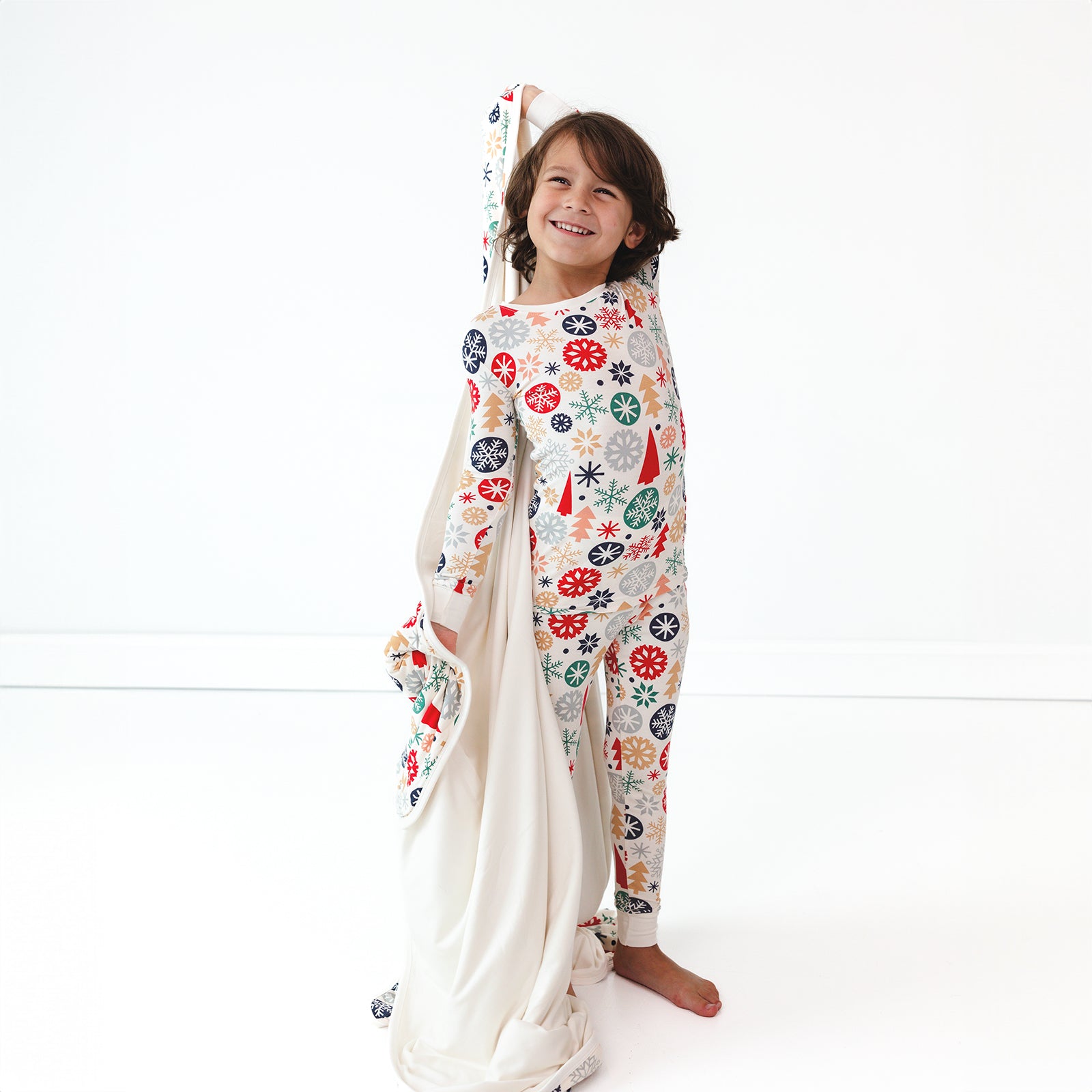 Smiling child posing in the Modern & Merry Two-piece Pajama Set while holding up the Modern & Merry Large Cloud Blanket® displaying the inside cream color