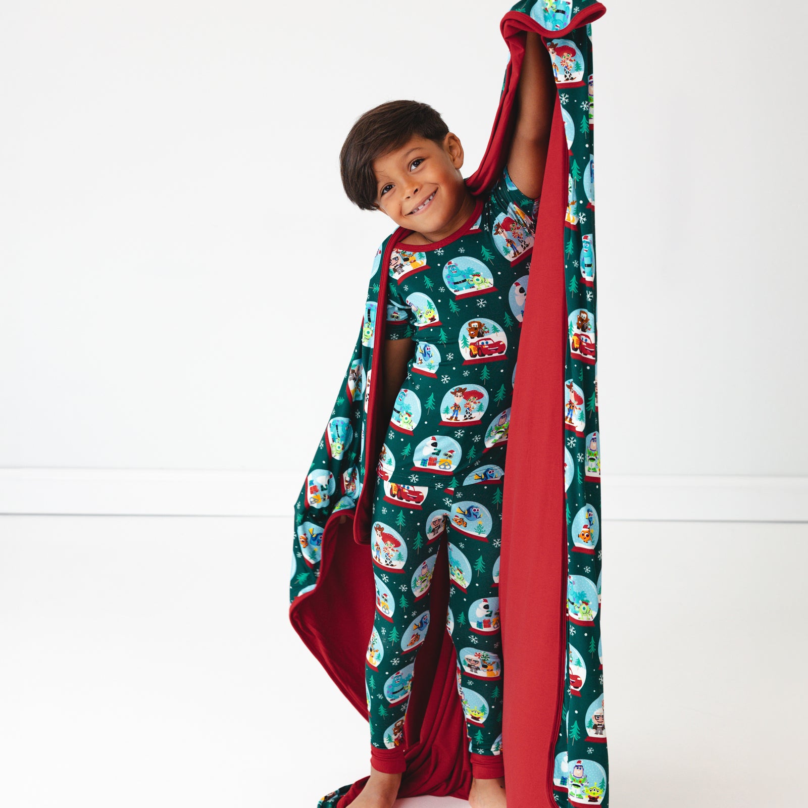 Image of a child wearing A Very Pixar Christmas Two-Piece Short Sleeve Pajama set and holding up a matching Large Cloud Blanket®