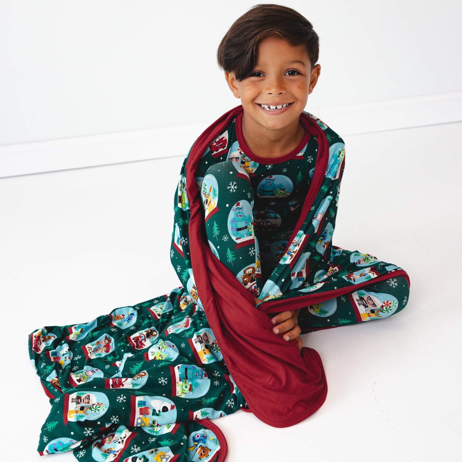 Image of a child sitting on the ground wrapped up in A Very Pixar Christmas Large Cloud Blanket® and wearing matching pajamas.