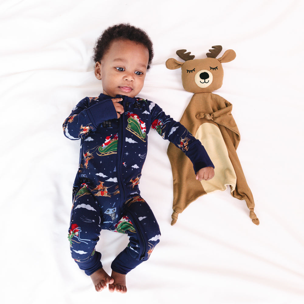 Alternative top view of baby laying down next to a Mistletoe the Reindeer Sleepyhead Lovey, while wearing the Santa's Sleigh Zippy