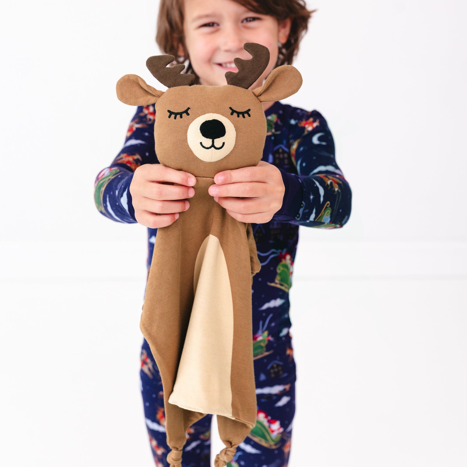 Close up image of child holding the Mistletoe the Reindeer Sleepyhead Lovey while wearing the Santa's Sleigh Two-piece Pajama Set